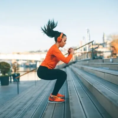 A young woman with a high ponytail and orange headphones performing jump squats on wide outdoor stairs.