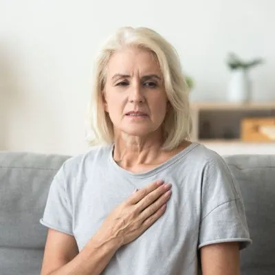 Older woman sitting on a couch with a concerned expression and her hand over her chest.