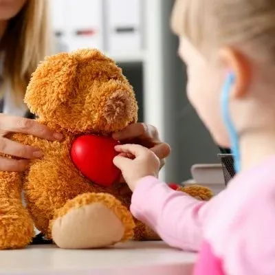 A child in a pink shirt using a blue toy stethoscope to listen to a red heart placed on a brown teddy bear.