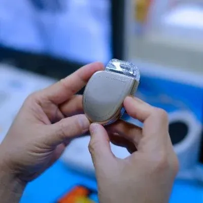 A person's hands holding a small, oval-shaped silver medical pacemaker.