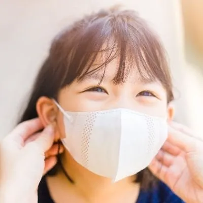 A smiling young girl with bangs wearing a white protective face mask.