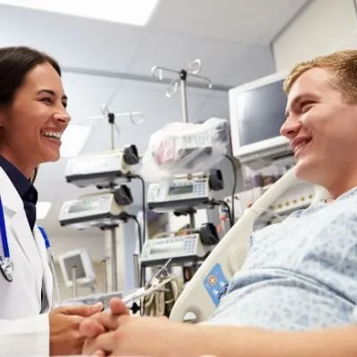 A smiling female doctor in a white lab coat and stethoscope talking to a male patient lying in a hospital bed.