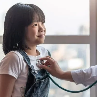 A young girl with dark hair wearing denim overalls and a white t-shirt smiles as a doctor uses a stethoscope to listen to her chest.