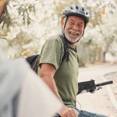 A smiling senior man with a grey beard wearing a bicycle helmet, green t-shirt, and backpack while sitting on his bike.