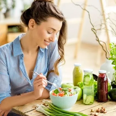 A smiling woman with her hair in a bun wearing a blue striped shirt sits at a wooden table, about to eat a fresh vegetable salad from a light blue bowl.