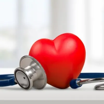 A red heart-shaped model resting on a white surface with a blue stethoscope placed against it.